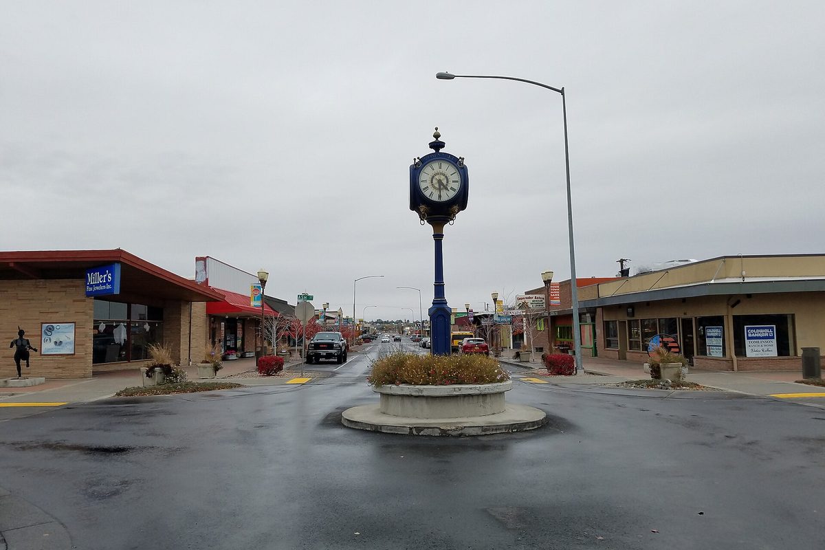 A blue post clock in a planted median on 3rd Avenue in downtown Moses Lake, with shops on both sides of a wet street under a gray sky
