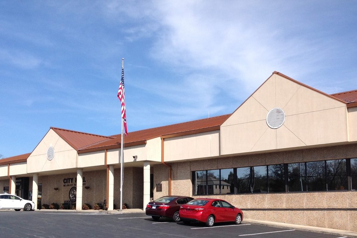 Mount Juliet City Hall, a long tan building with a red metal roof and a flagpole, with two red sedans parked out front under a blue sky