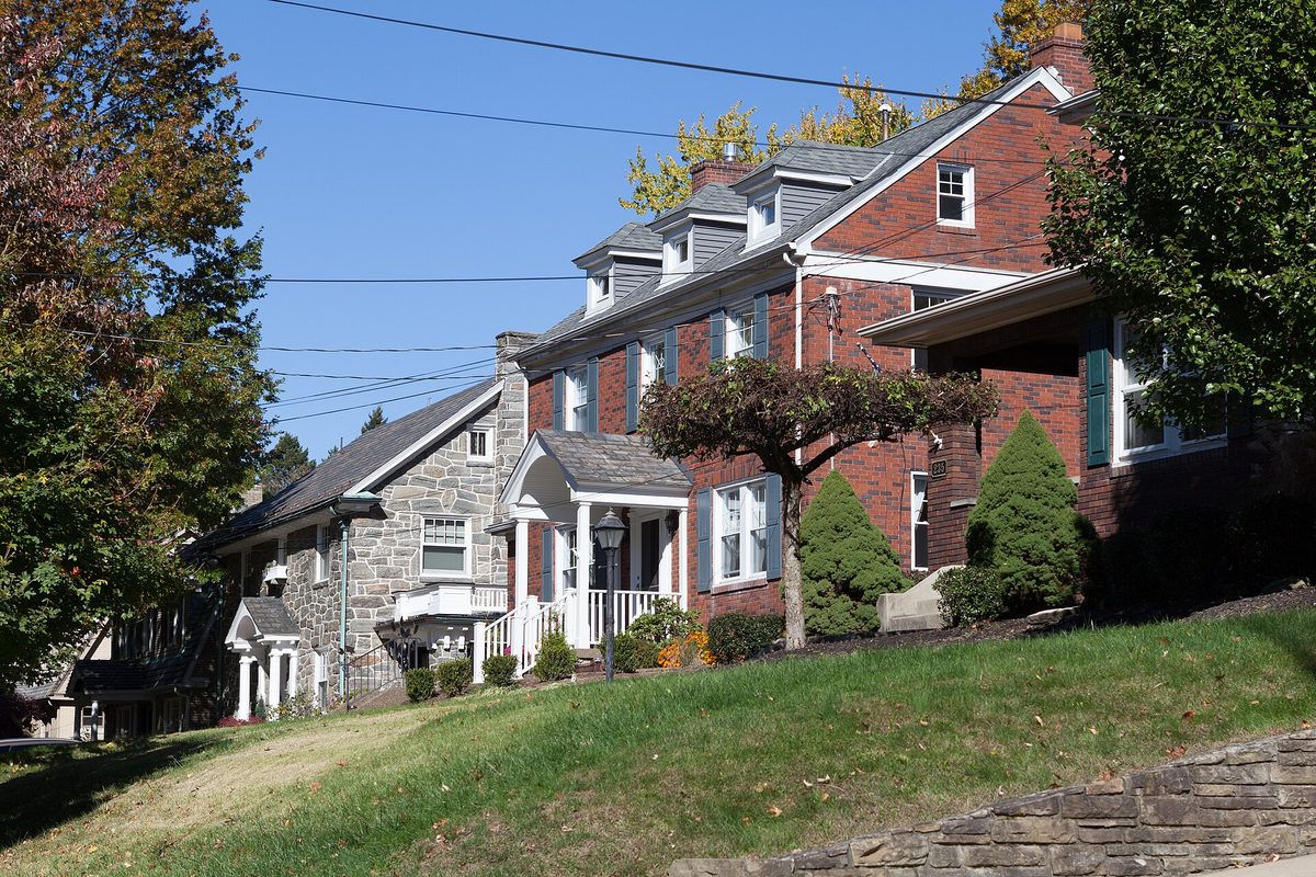 Brick and stone two-story houses with dormers and a white front porch on a grassy rise in the Mt. Lebanon Historic District, autumn trees around them