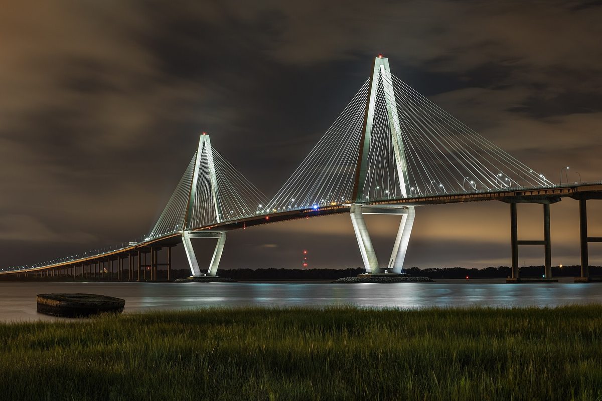 The Ravenel Bridge lit up at night over dark water, seen across marsh grass from Mount Pleasant