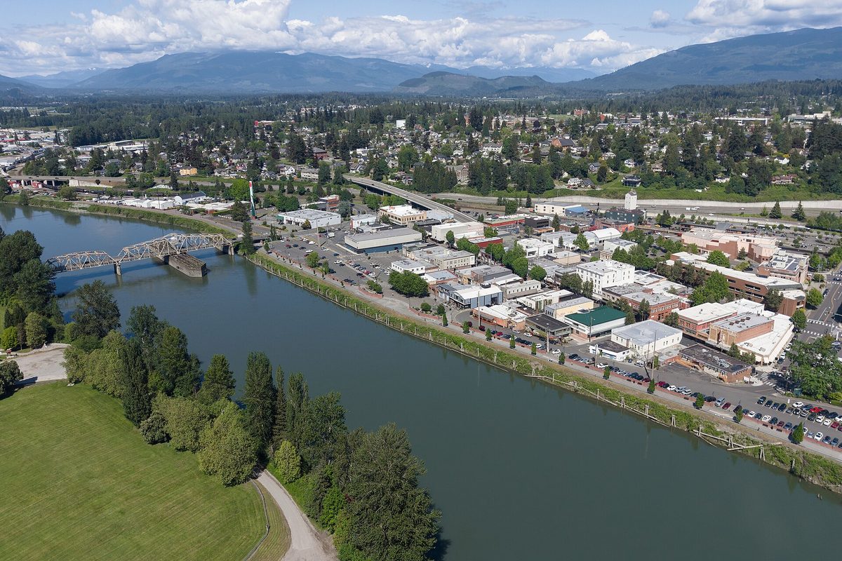 Aerial view of downtown Mount Vernon beside a wide river, with a steel truss bridge, a highway bridge and forested mountains behind