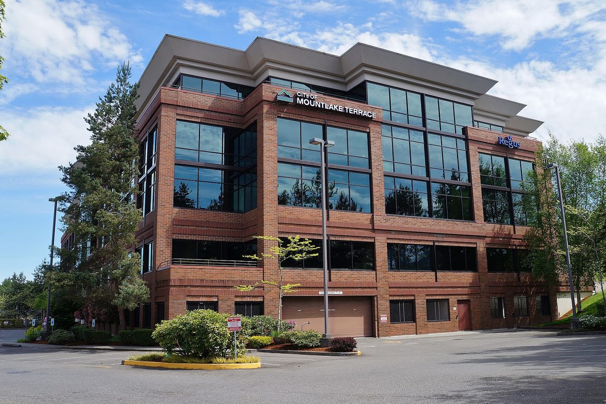 Four-story brick and glass office building with a City of Mountlake Terrace sign above the entrance, a landscaped island and parking lot in front