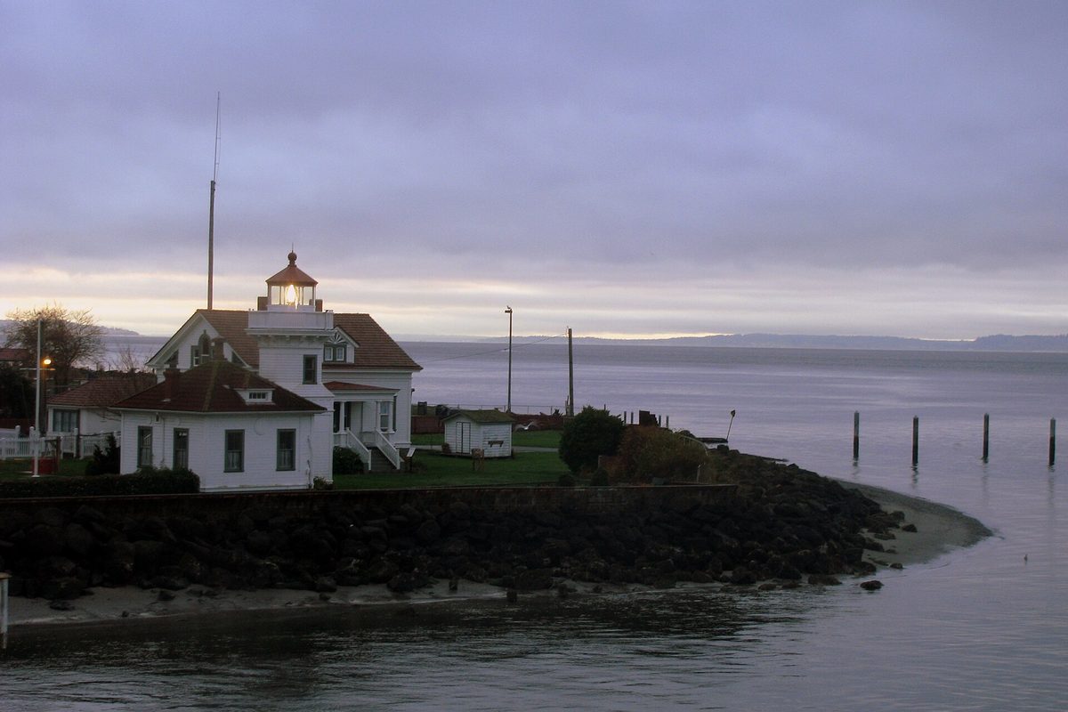 The white Mukilteo Light with its red roofs on a rock seawall at dusk, with Puget Sound and distant hills behind