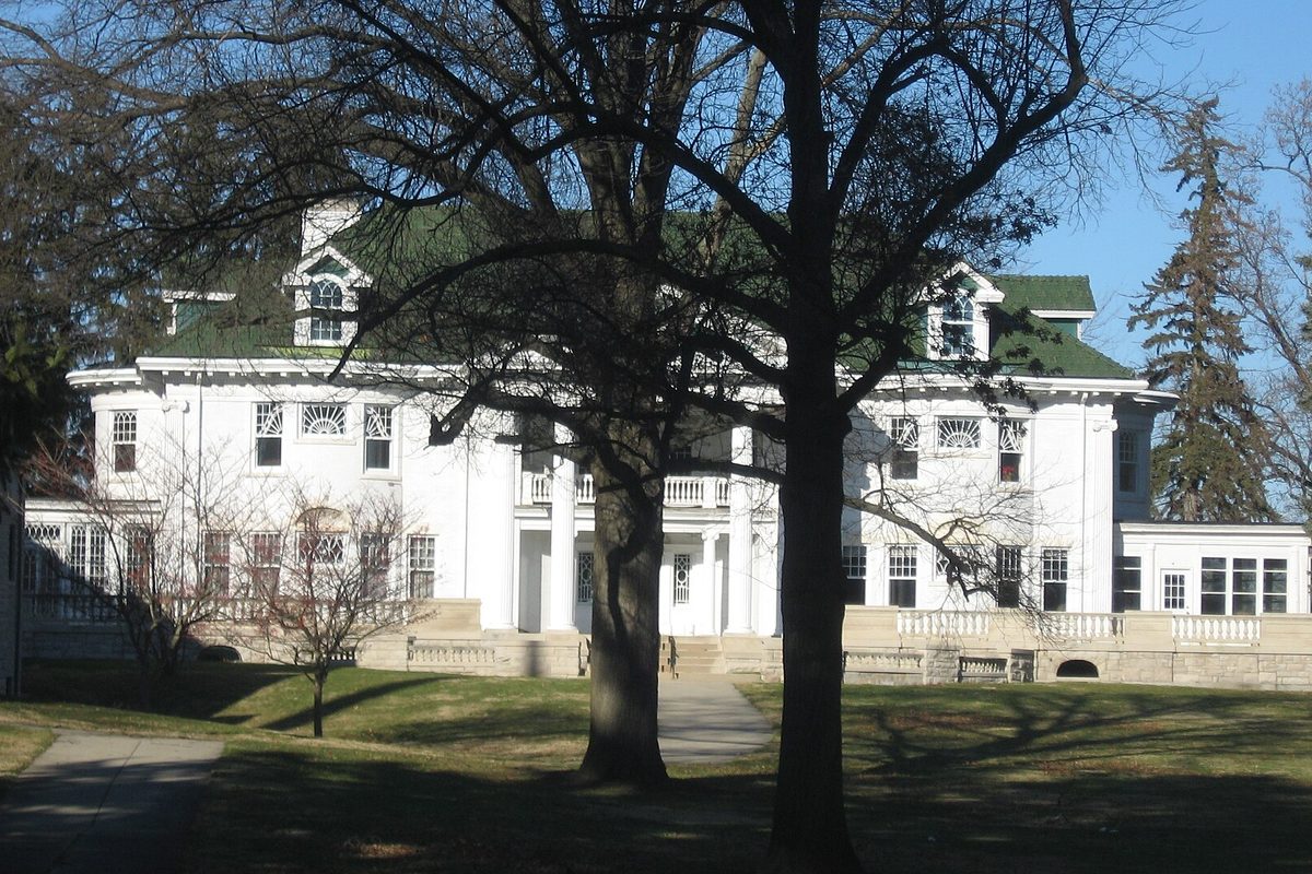The white Alva Kitselman House in Muncie, with a green roof and a columned porch, seen through bare trees across a lawn