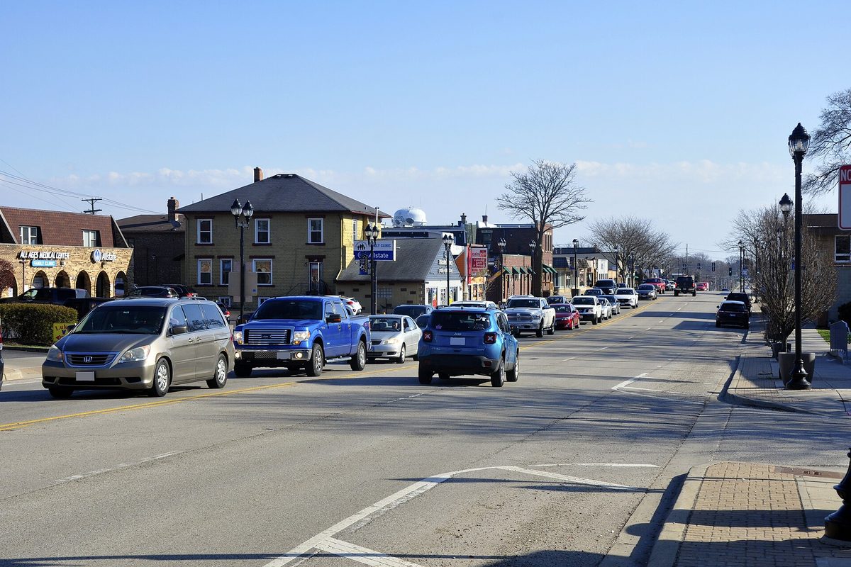 Cars and pickups driving along a downtown Mundelein street lined with two-story shops and black lamp posts on a sunny day