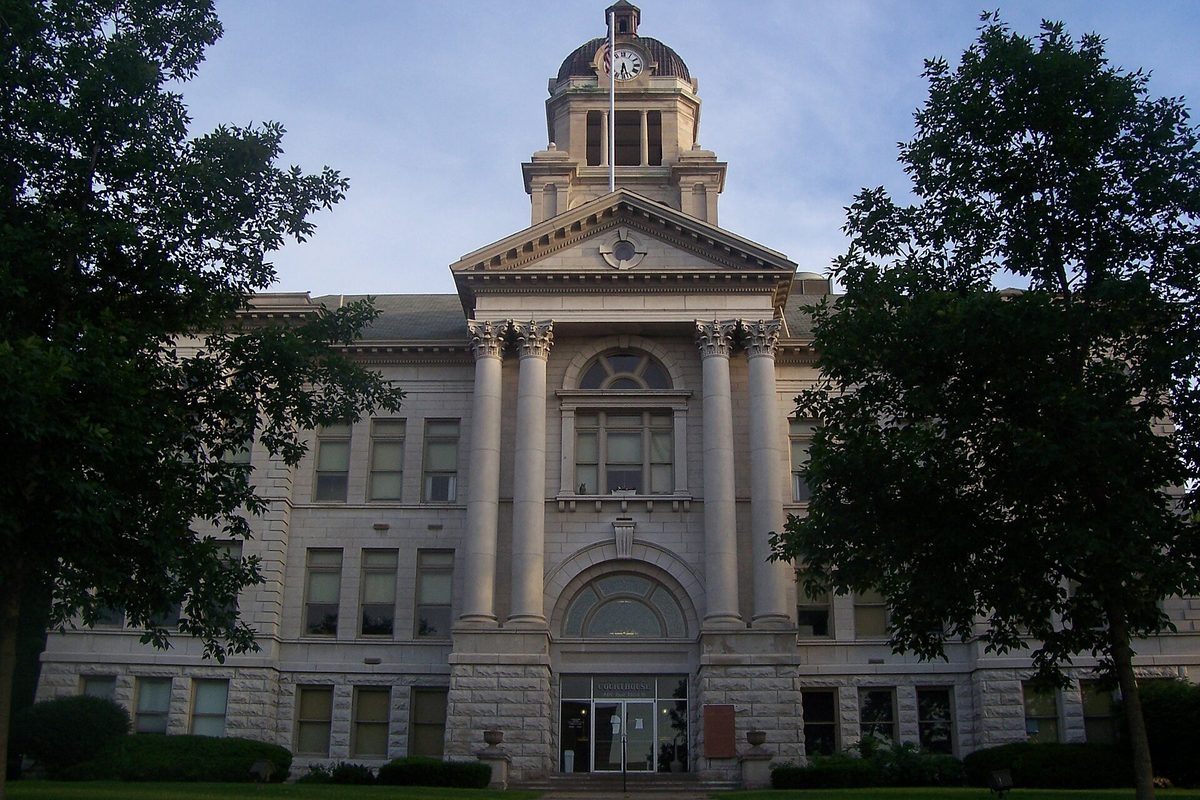 The domed stone Muscatine County Courthouse, with columns and a clock tower, framed by two large trees at dusk