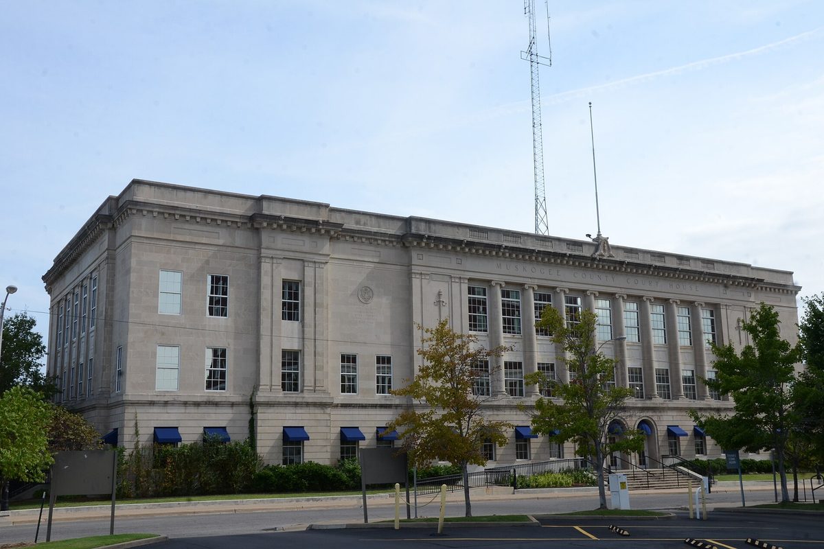 The Muskogee County Courthouse, a pale stone building with tall windows, blue awnings along the ground floor and a radio mast rising behind it