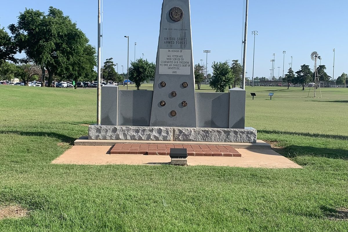 A granite veterans memorial obelisk on a stone and brick base in Wild Horse Park, Mustang, with a windmill and ball-field lights behind it