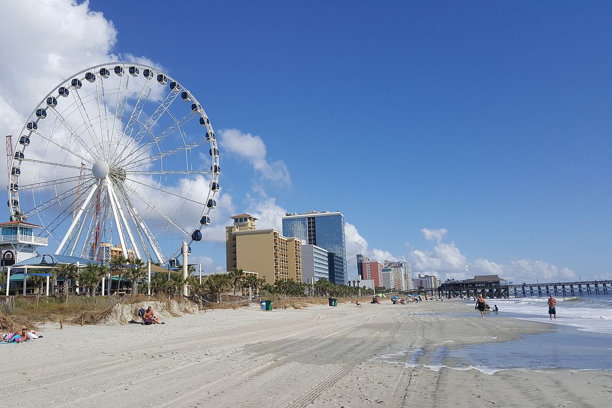 A large white observation wheel on the sand at Myrtle Beach, with high-rise hotels behind it and a fishing pier reaching into the surf