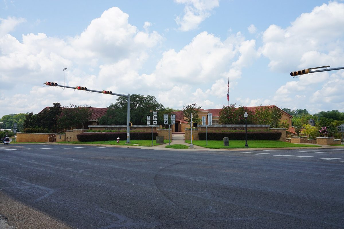 The low red-brick Nacogdoches County Courthouse with a flagpole and hedges, seen across a wide intersection under traffic signals and white clouds