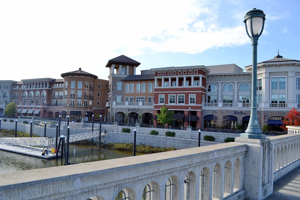 Brick and stucco buildings with arched windows and a clock tower along the Napa River, seen past a stone bridge railing and a green lamppost