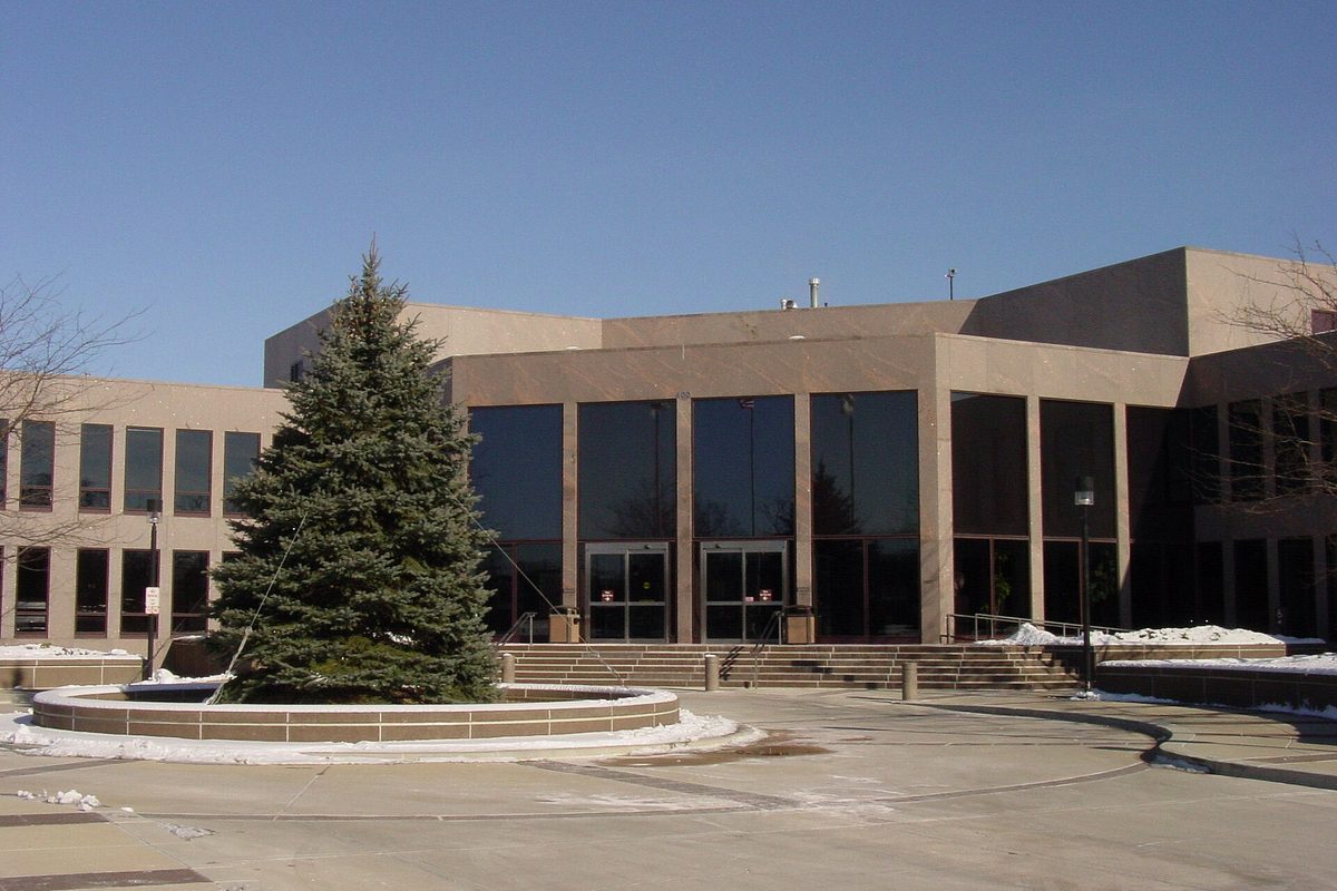 Main entrance of Naperville City Hall, a tan building with tall dark windows, a spruce tree in a round planter and snow on the ground
