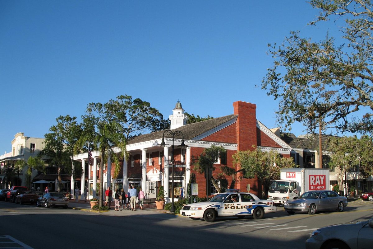 A red-brick building with white columns and a white cupola on a sunny Naples street corner, with people on the sidewalk, palm trees and a police car passing