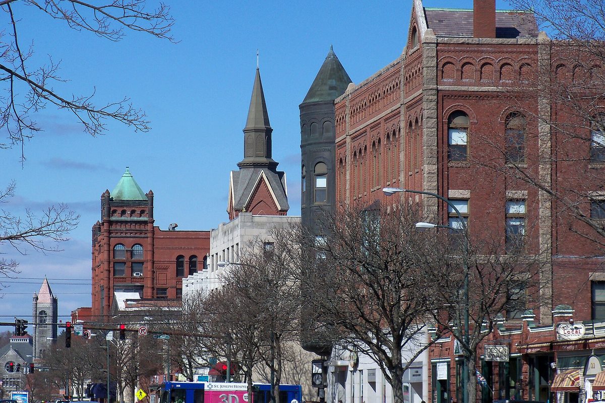 Red-brick Victorian buildings, a pointed church steeple and a green-domed tower along Main Street in Nashua, with bare trees and a passing bus