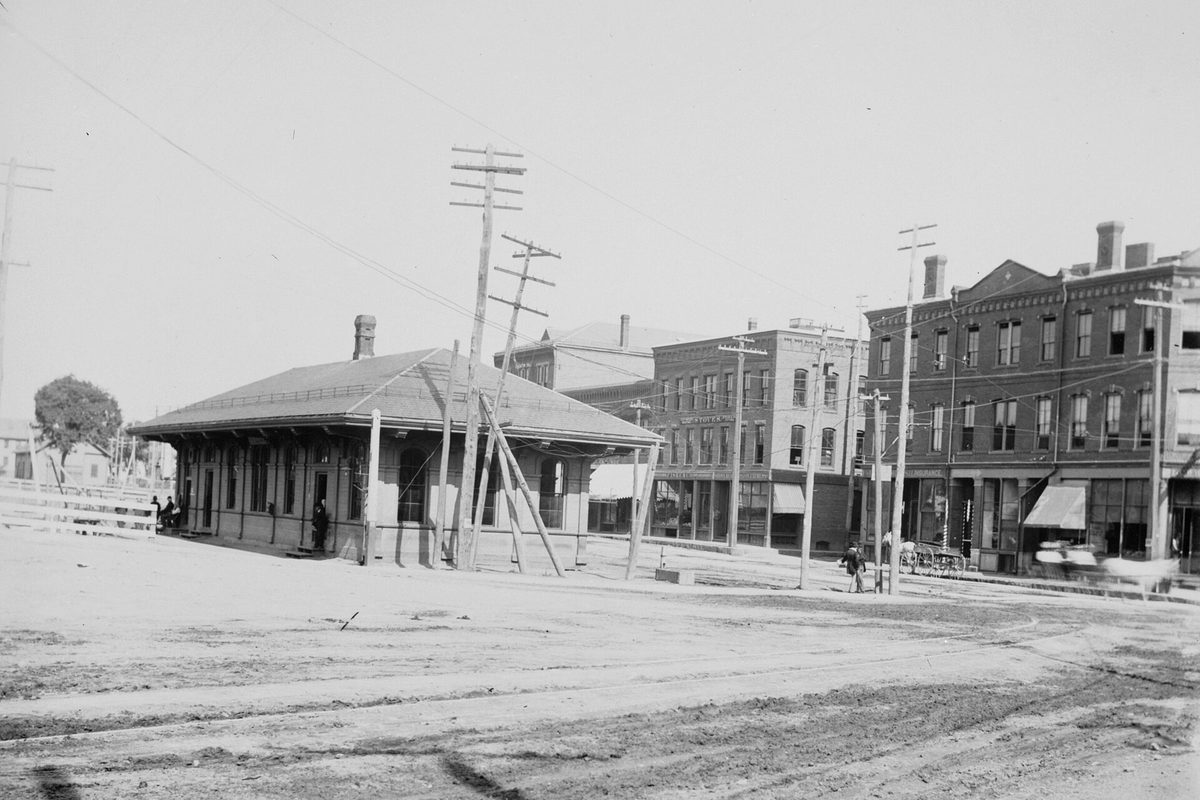 Black-and-white 1890s photo of the old Natick station, a low wooden depot on a dirt street lined with telegraph poles and brick commercial blocks