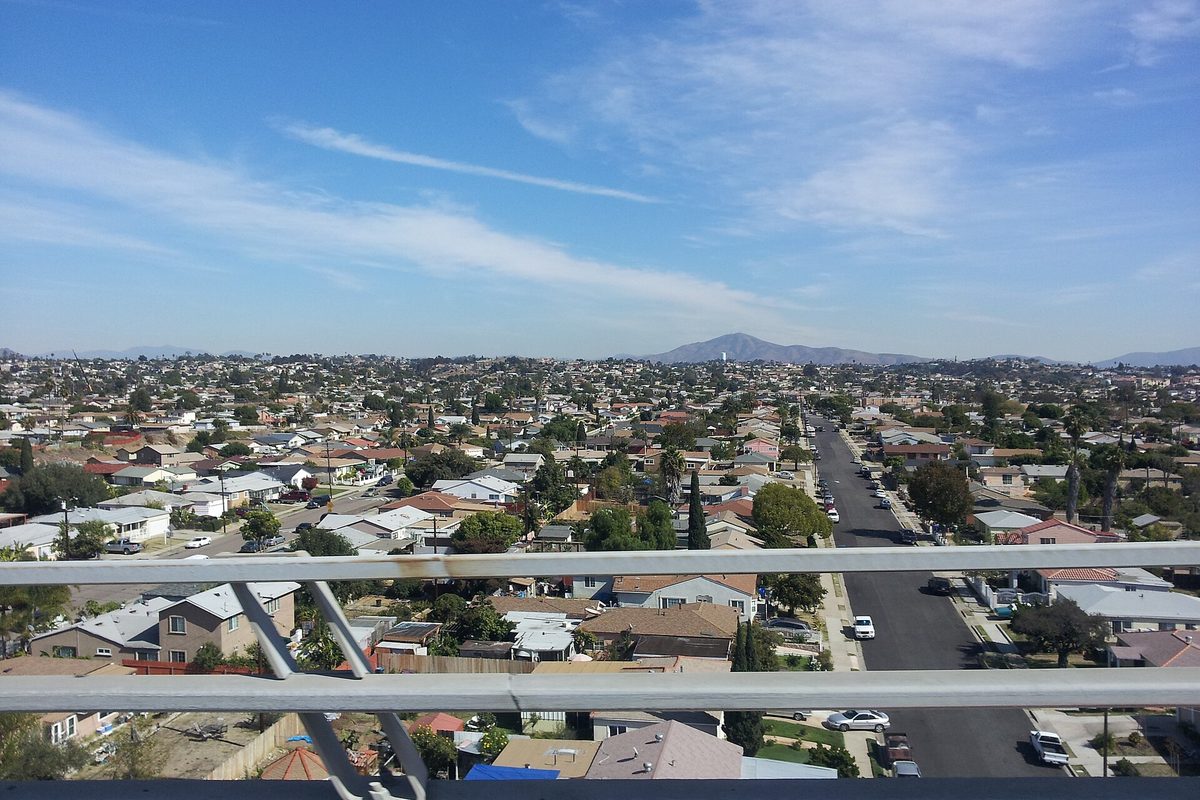 Rooftops of a National City neighborhood seen through a freeway overpass railing, with a mountain on the horizon under a blue sky