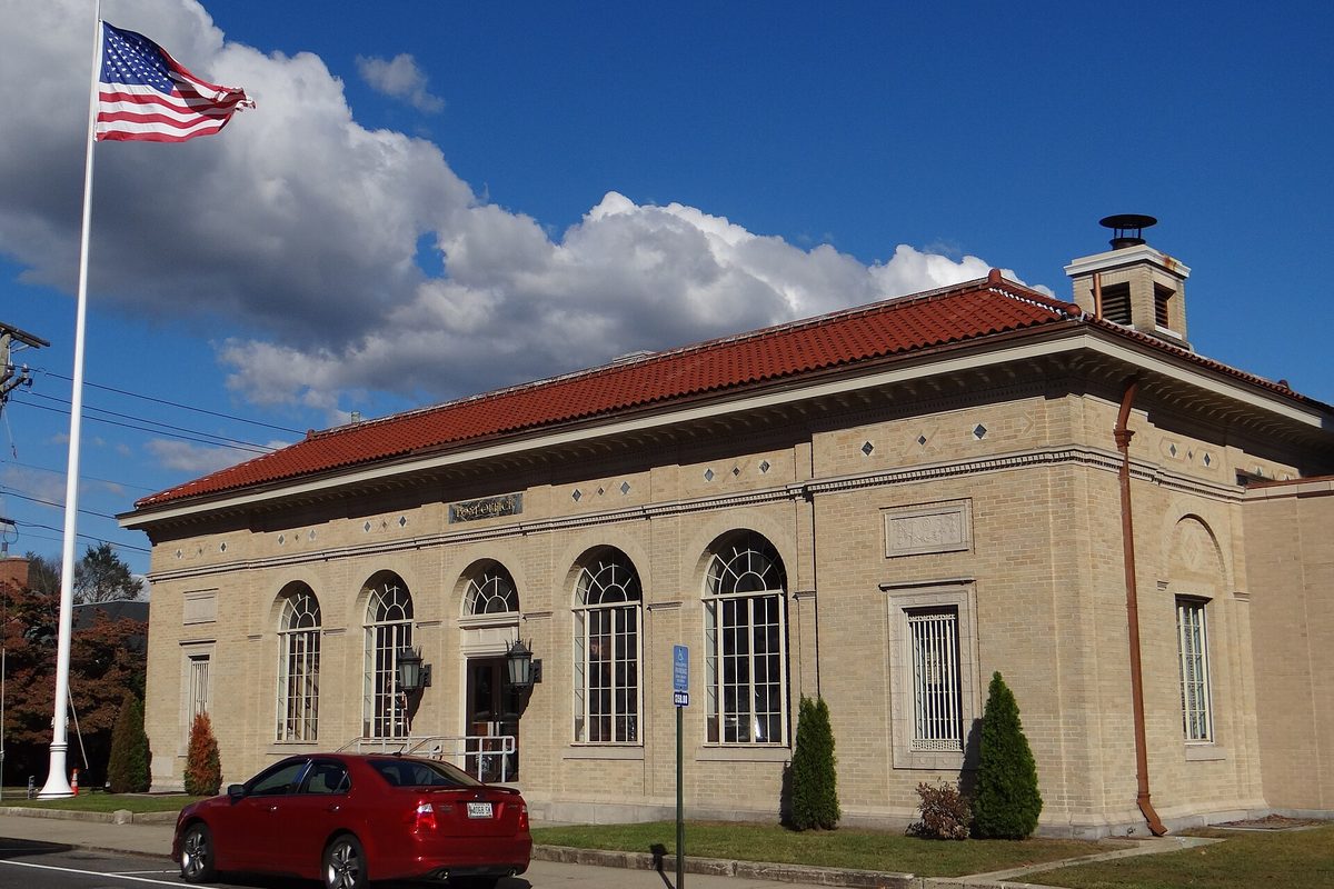 Naugatuck's main post office, a tan brick building with a red tile roof and tall arched windows, an American flag on a pole and a red car parked at the curb