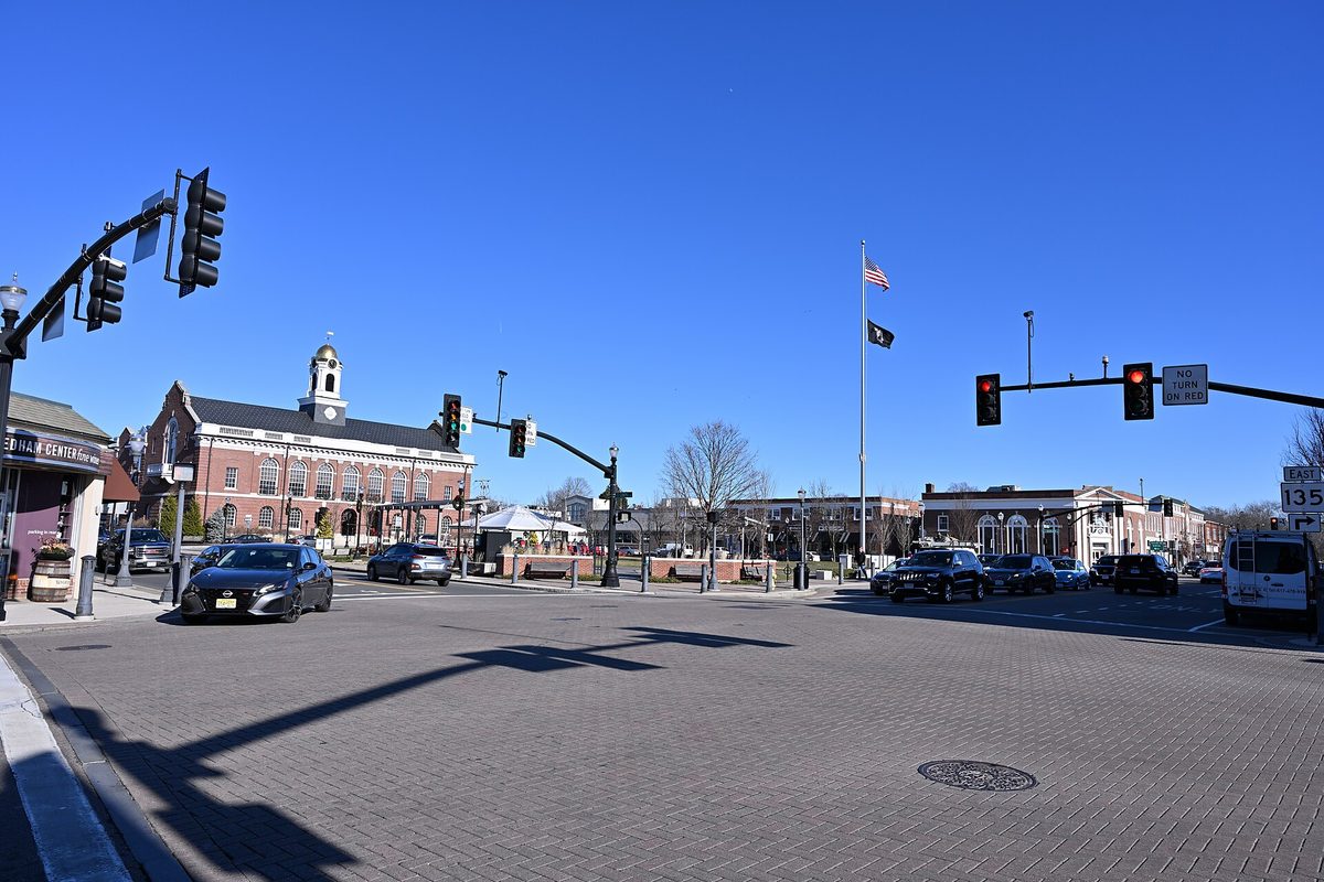 The crossroads in Needham Center under a clear blue sky, with traffic lights, cars at the intersection and the brick town hall with a white cupola on the left