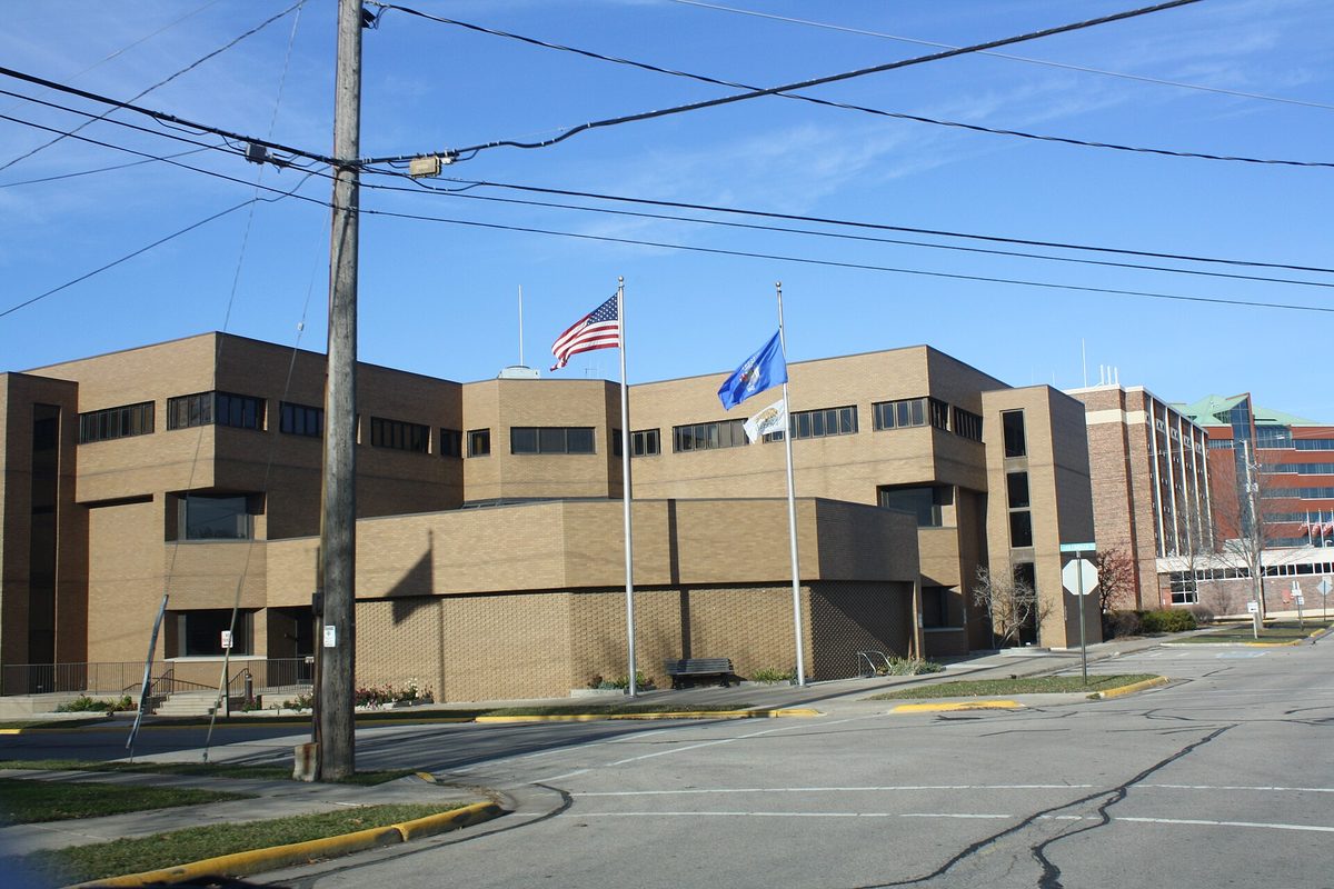 Neenah City Hall, a tan brick building with long rows of dark windows, with the U.S. and Wisconsin flags flying out front under a blue sky