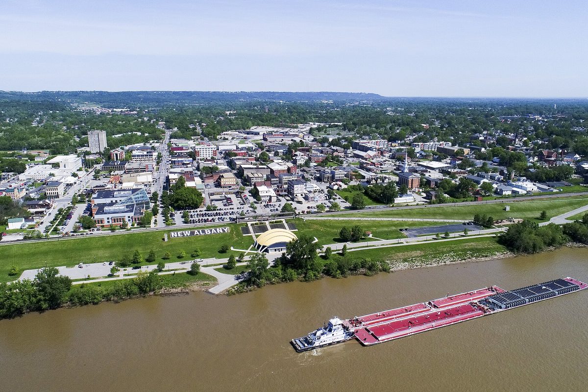 Aerial view of New Albany from the Ohio River, with a towboat pushing barges past the riverfront lawn and a large NEW ALBANY sign