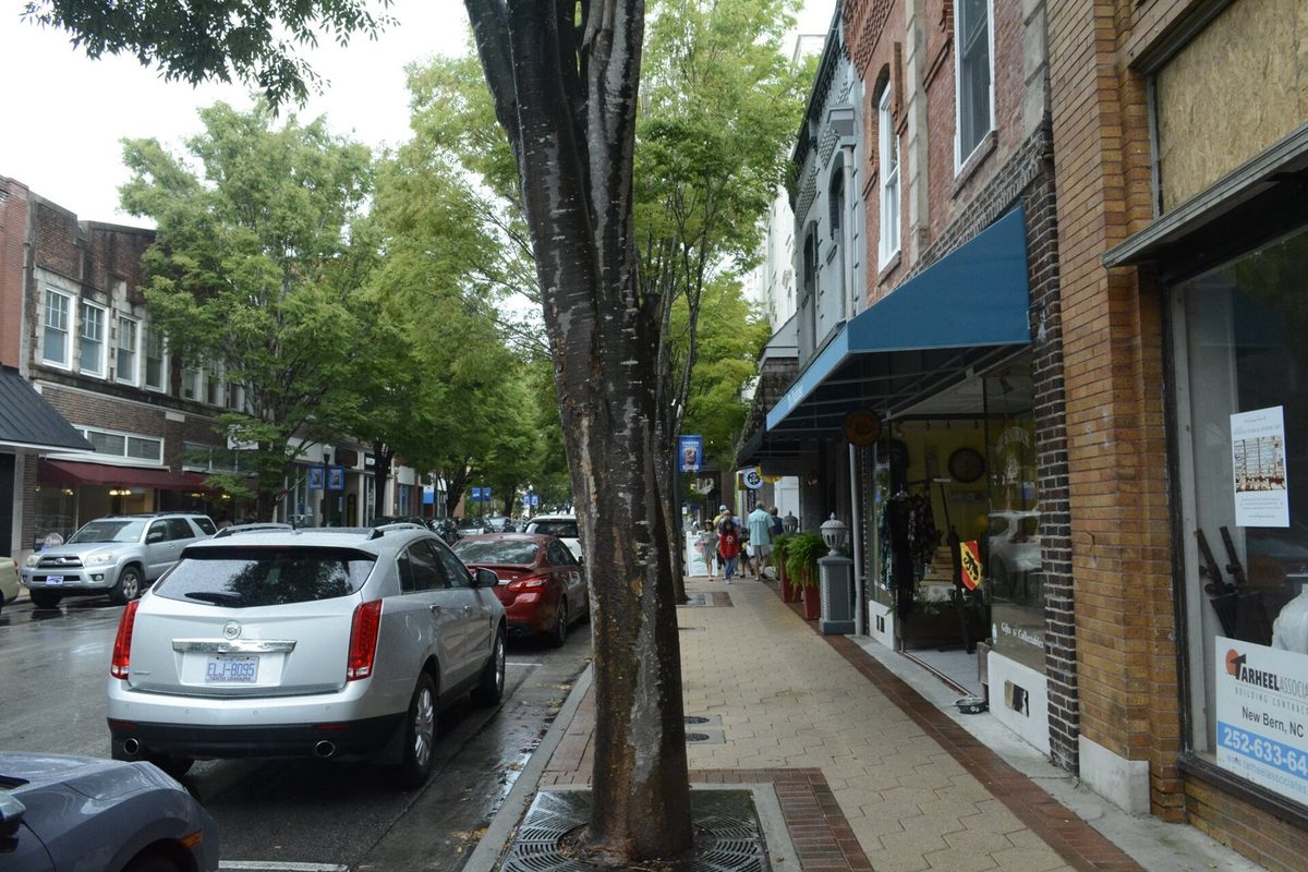 Middle Street in downtown New Bern after rain, with brick storefronts and a blue awning on the right and cars parked under shade trees