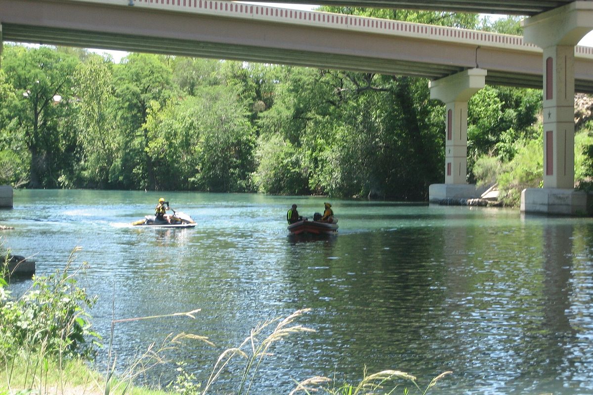 The green Guadalupe River in New Braunfels flowing under a concrete highway bridge, with two small boats carrying people in life jackets and trees along the far bank