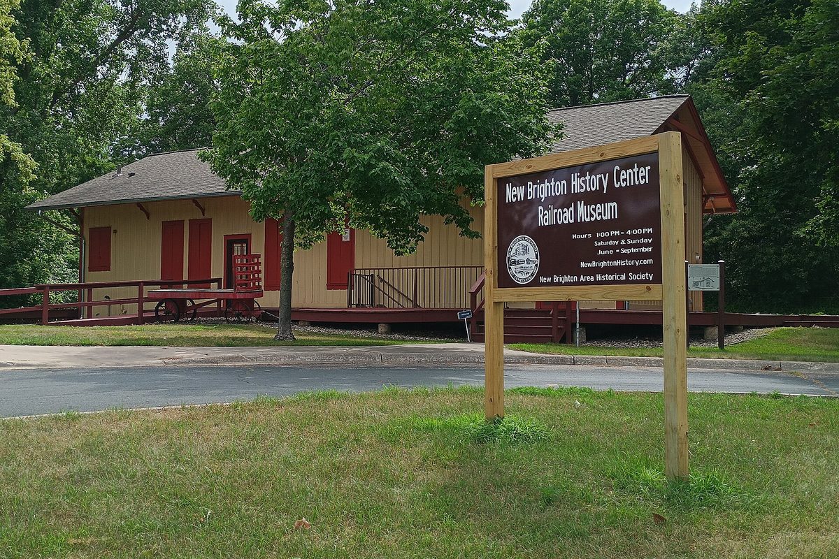 The yellow New Brighton History Center depot with red doors and a wooden deck, behind a sign for the Railroad Museum in Long Lake Regional Park