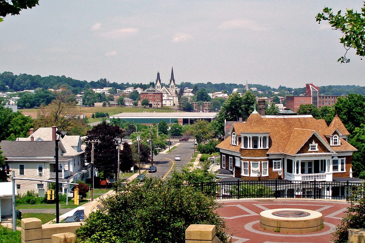 View north from Walnut Hill Park in New Britain, past a brick plaza and a shingled Victorian house to a street, rooftops and twin church spires