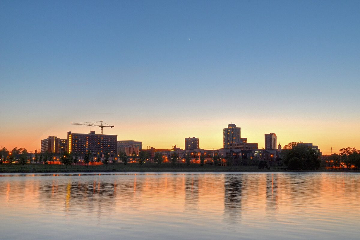 The New Brunswick skyline at sunset, with office towers, apartment blocks and a construction crane reflected in calm water under an orange sky