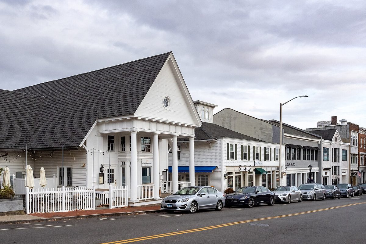 A white building with a columned porch and a row of shops on a downtown New Canaan street, cars parked along the curb under a gray sky