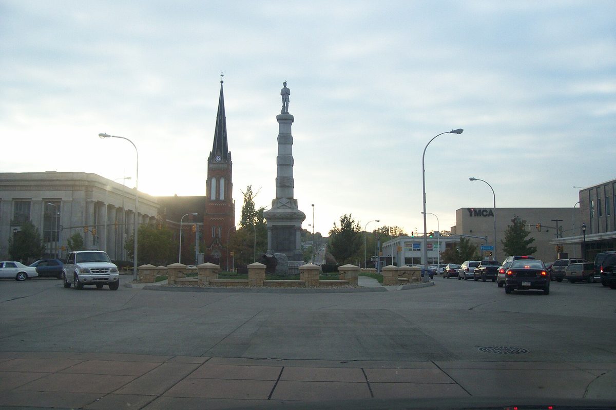 Kennedy Square in downtown New Castle at dusk, a tall monument topped by a statue in a traffic circle, a brick church spire to the left and cars passing
