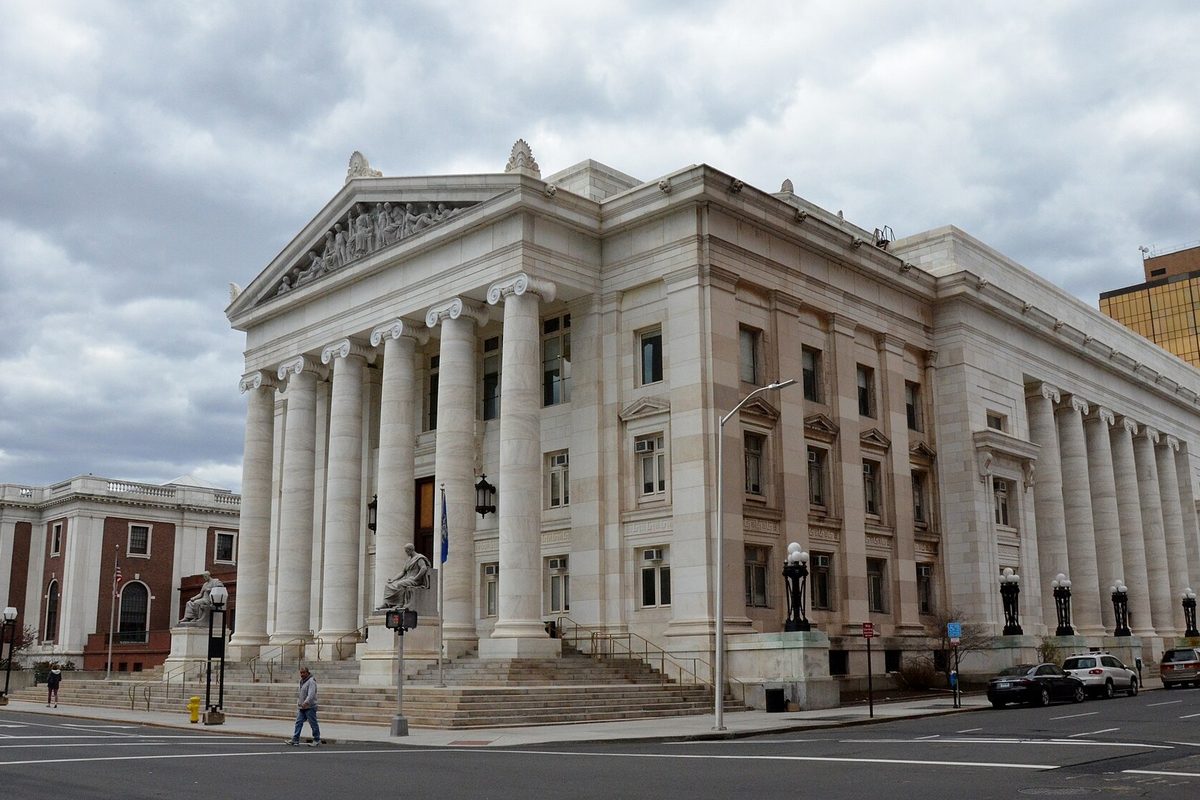 A white marble building with tall columns and a sculpted pediment on a downtown New Haven street corner, a person walking past and cars parked along the curb under a cloudy sky