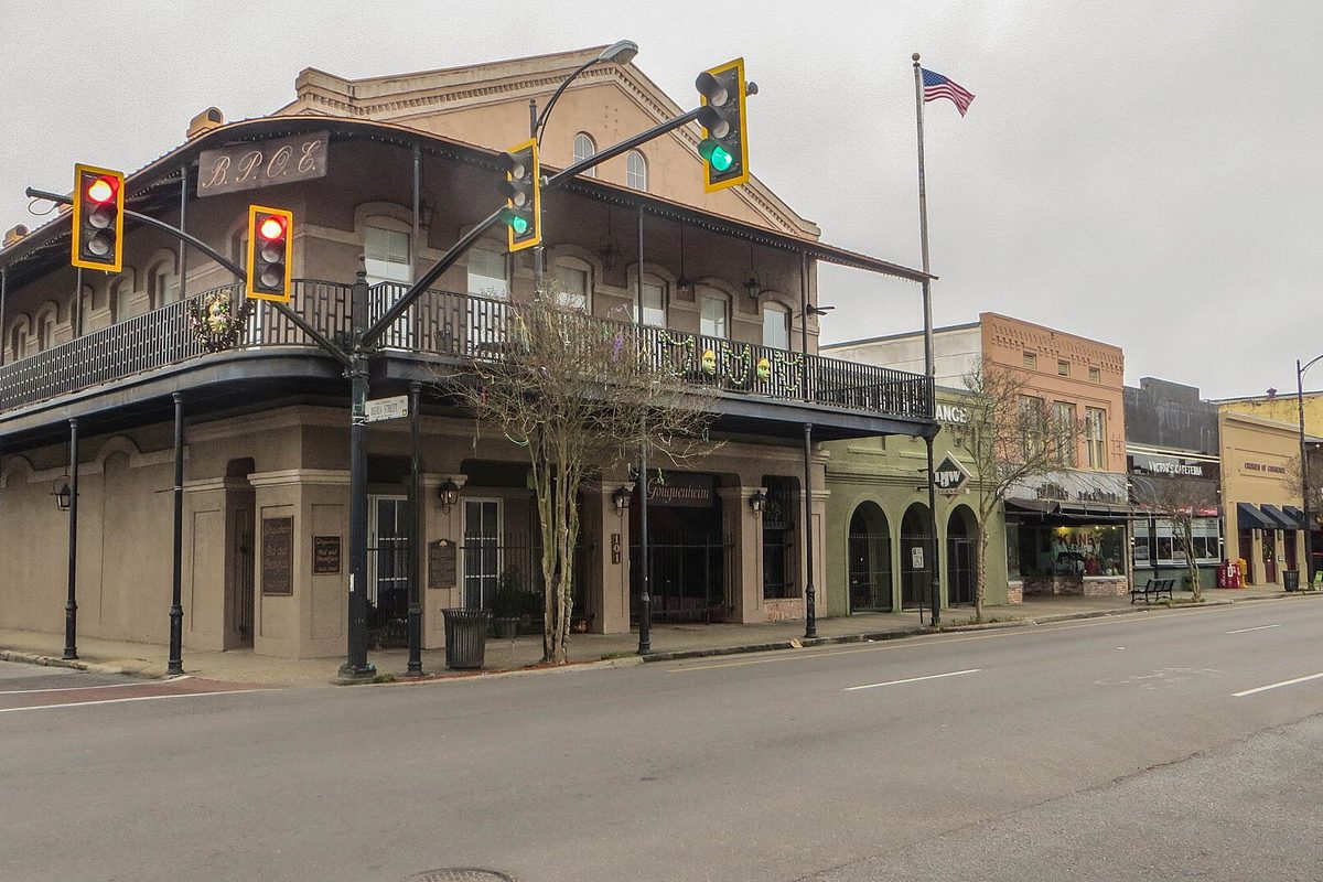 A two-story downtown New Iberia corner building with an iron balcony, traffic lights and a U.S. flag under a gray sky