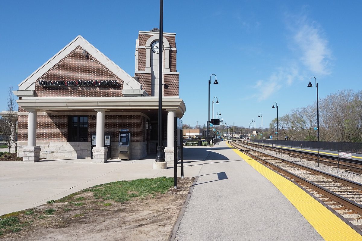 The red brick New Lenox Metra station with a clock tower and white columns beside the inbound platform and tracks, under a clear sky