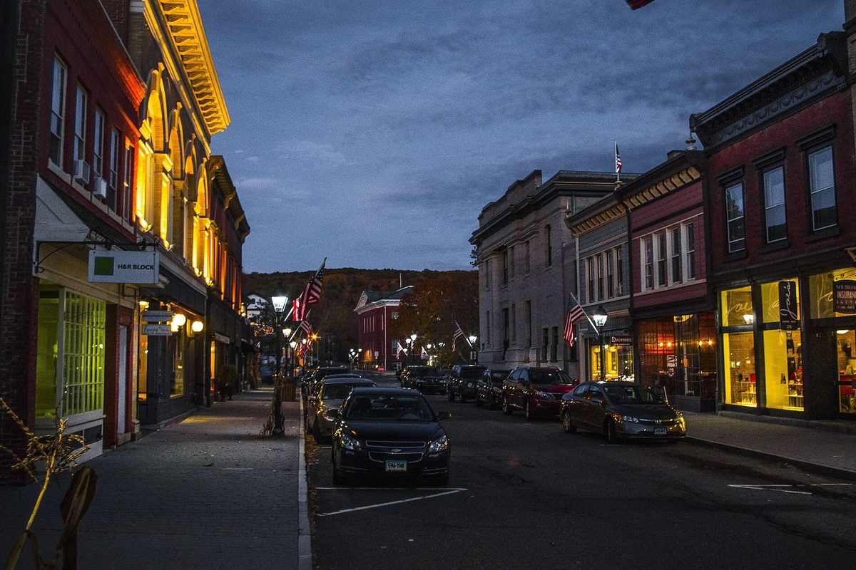 Bank Street in New Milford at blue hour: lit storefronts, American flags on brick buildings and cars parked along the curb