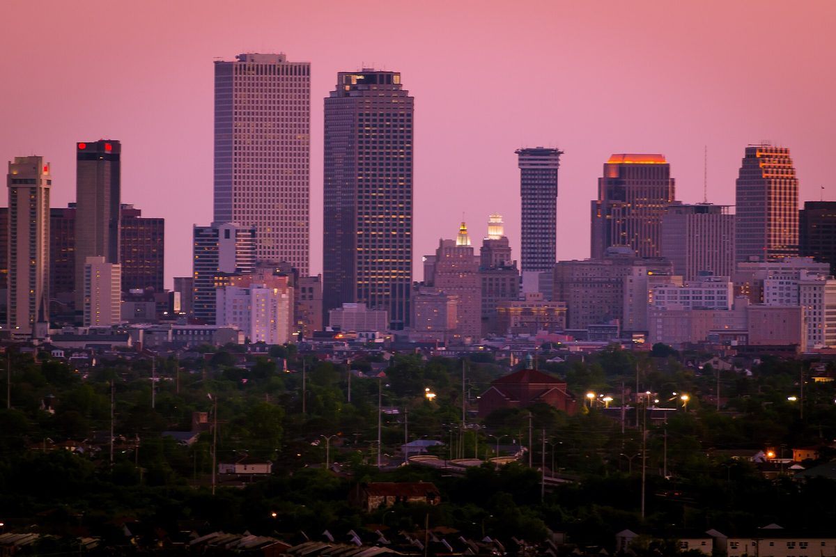 The downtown New Orleans skyline at dusk under a pink sky, with streetlights and tree-lined neighborhoods in the foreground