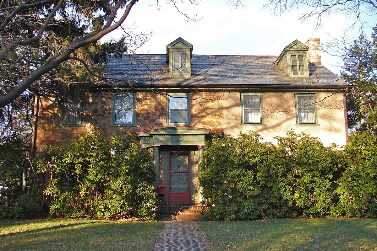 The stone Anderson House in Newark, a two-story historic home with dormer windows, a slate roof and a red front door behind large green shrubs