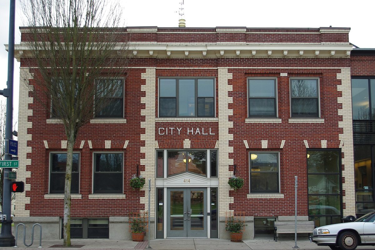 Newberg's red brick City Hall with City Hall lettered above the entrance, on a street corner beside a First Street sign