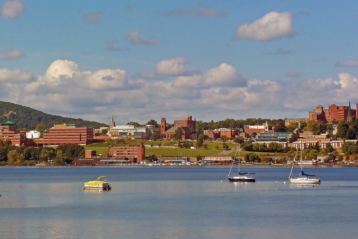 Downtown Newburgh's brick buildings climbing the hillside above the Hudson River, with sailboats and a small pontoon boat moored in the foreground