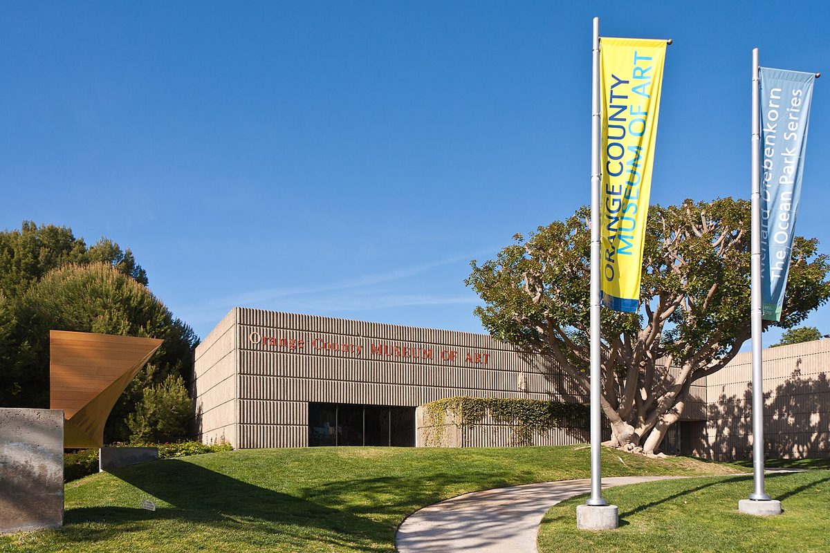 The ribbed stone front of the Orange County Museum of Art with yellow and blue banners on tall poles, a curving path and a large tree