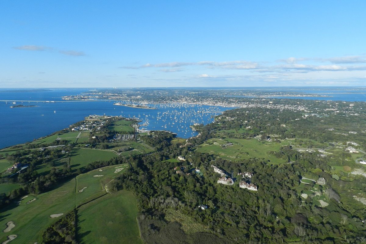 Aerial view of Newport, Rhode Island, with a harbor full of moored boats, a long bridge across the bay in the distance and green golf courses and large houses in the foreground