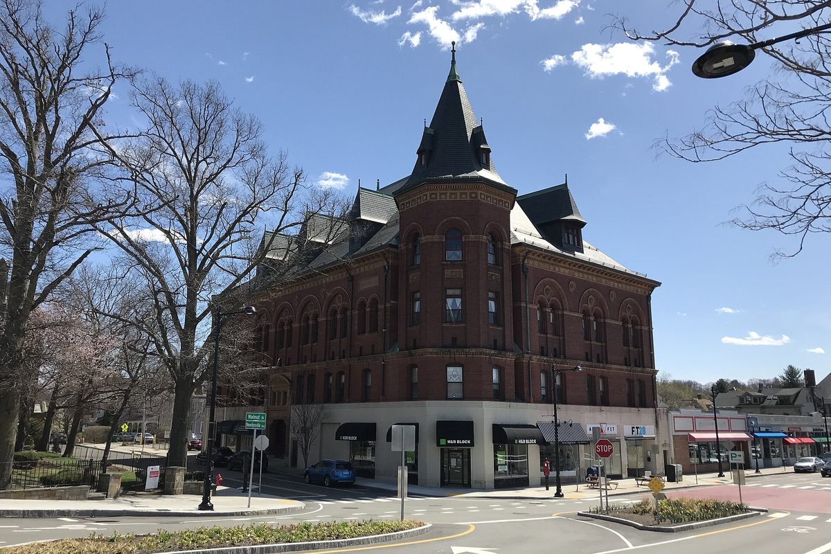 The red brick Masonic Building in Newtonville with a pointed slate turret, shops at street level and bare trees on a sunny spring day