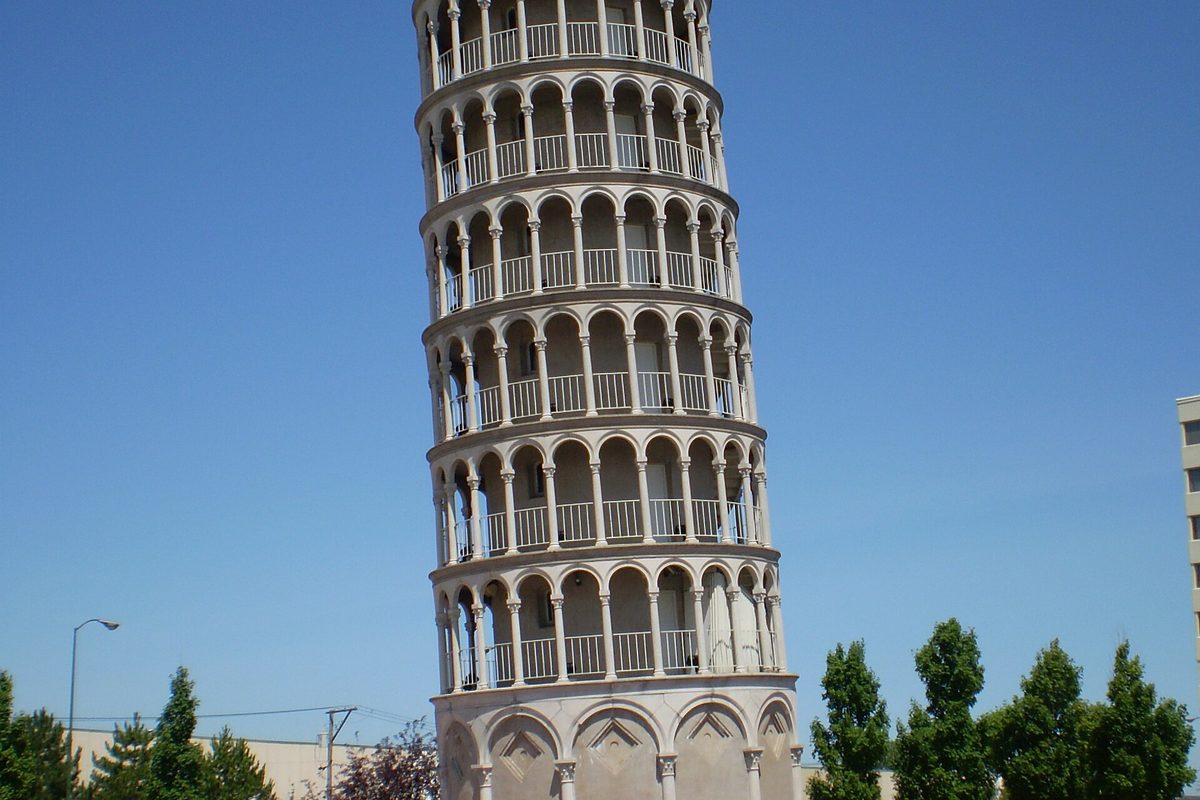 The Leaning Tower of Niles, a white tower of stacked arched balconies, tilting against a clear blue sky with trees at its base