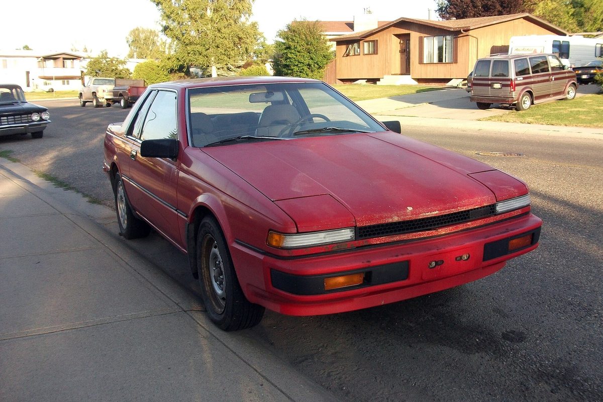 A faded red 1980s Nissan 200SX coupe parked at a curb on a residential street, with an old van and houses behind it