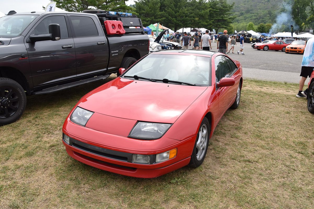 A red Z32 Nissan 300ZX parked on the grass at a car show, beside a dark gray pickup truck