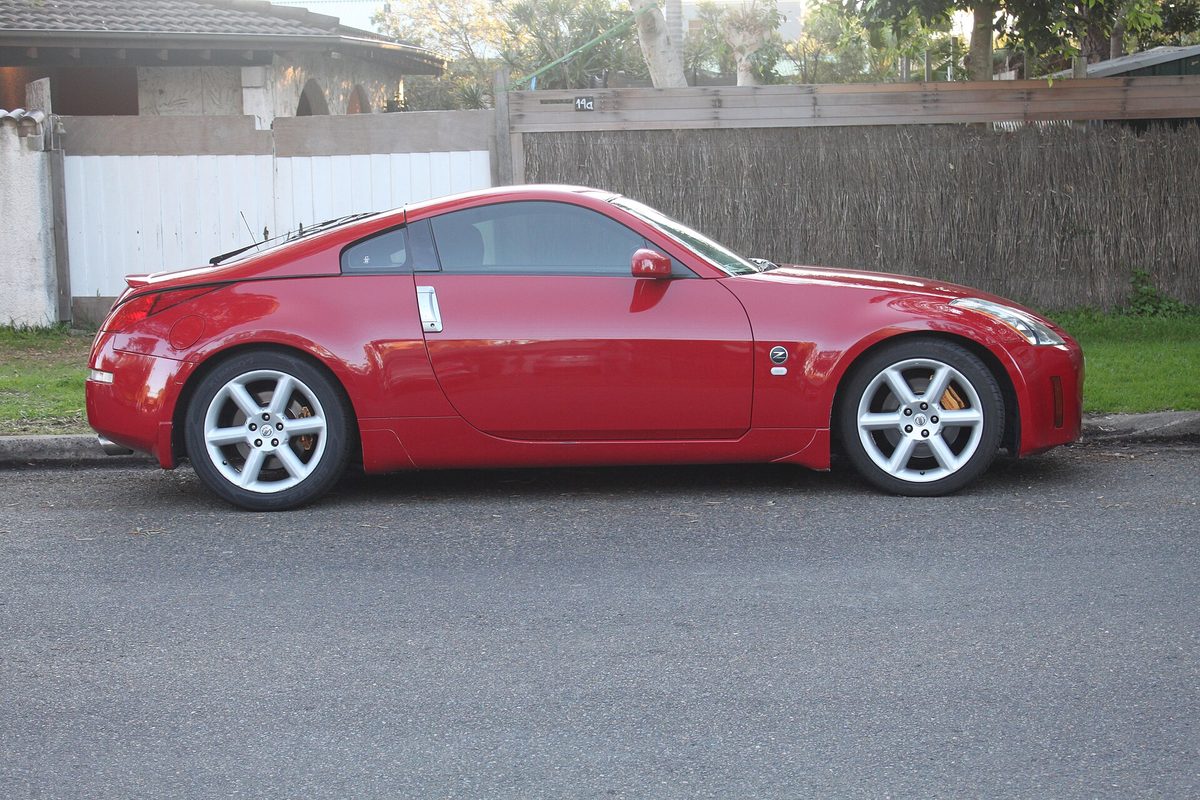 A red 2003 Nissan 350Z coupe seen from the side, parked at a curb in front of a wooden fence