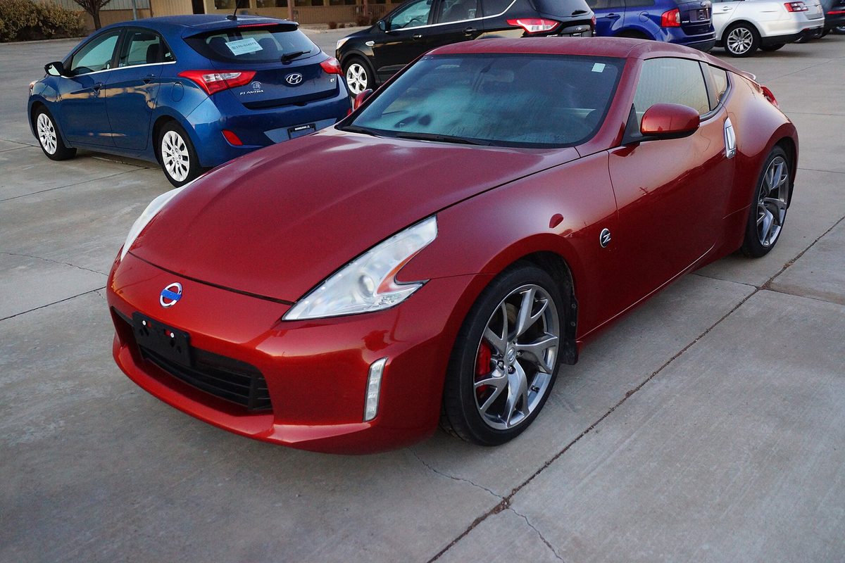 A red 2014 Nissan 370Z coupe on a concrete lot, with a blue hatchback parked behind it