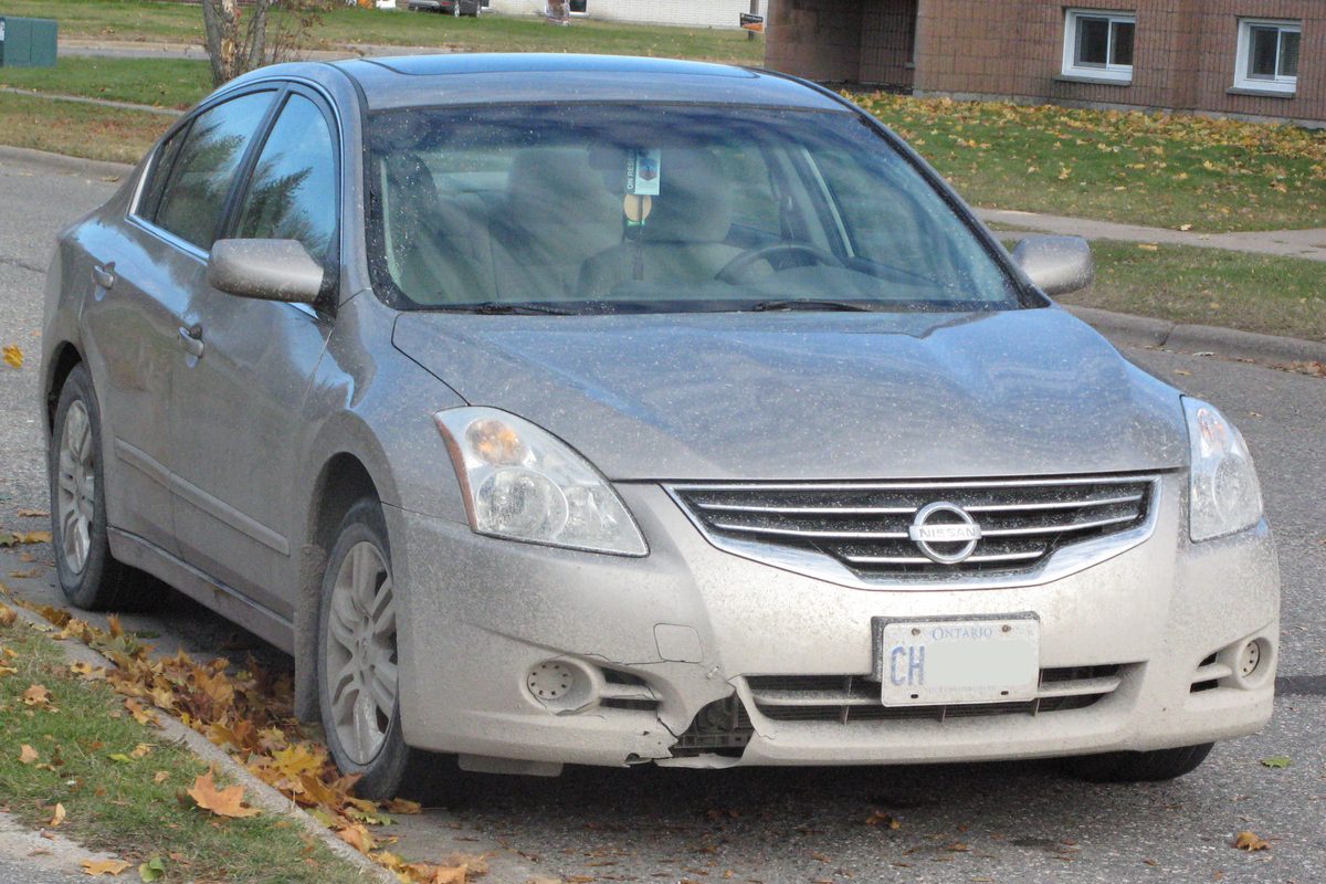 A dusty silver 2012 Nissan Altima with a cracked front bumper, parked at a curb in fallen leaves
