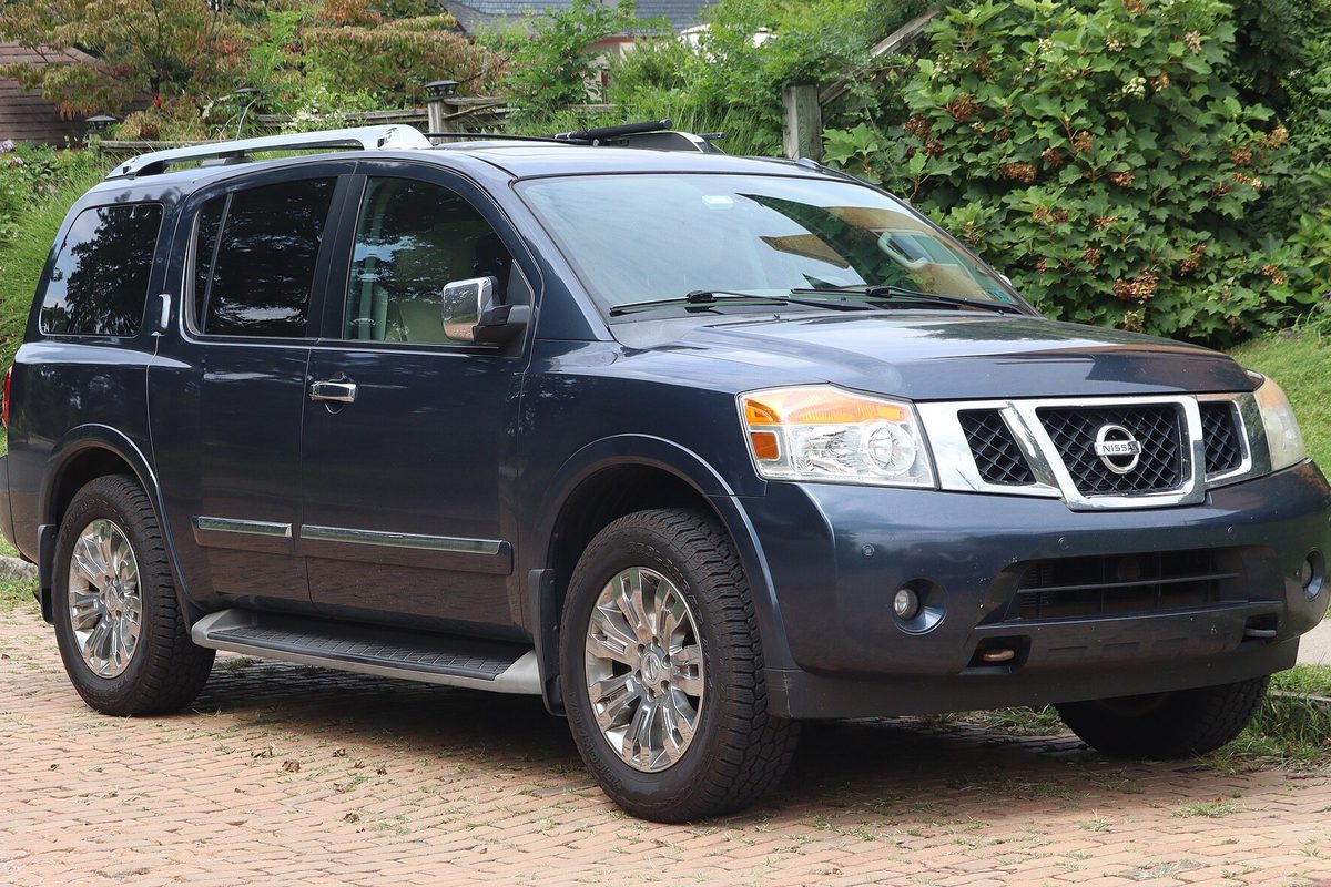 A dark blue 2015 Nissan Armada Platinum with chrome wheels and roof rails, parked on a brick driveway in front of tall hedges