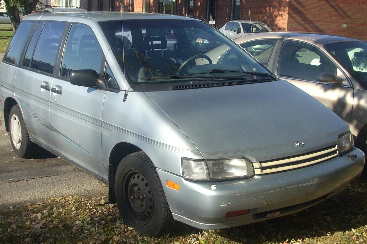 A pale blue-gray Nissan Axxess compact van with steel wheels and a scuffed front bumper, parked beside a tan sedan